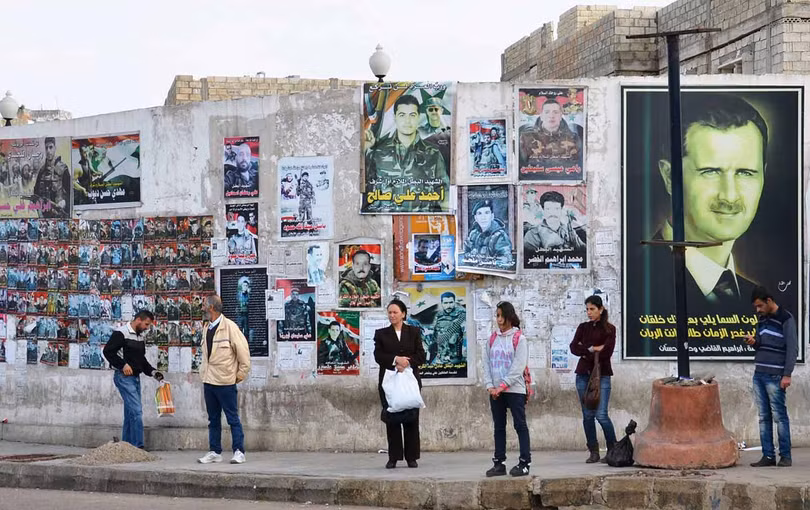 Người dân Syria chờ xe bus tại Tartus trước chân dung của tổng thống Bashar al-Assad cùng ảnh của những người đã hy sinh khi chiến đấu bảo vệ chế độ tại Syria.