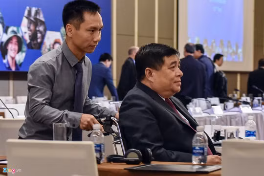 Bộ trưởng Nguyễn Chí Dũng phải ngồi xe lăn và nhờ người hỗ trợ khi đến dự diễn đàn. Ảnh: Việt Linh.