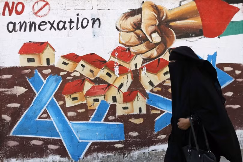 Một phụ nữ Palestine đi qua bức tranh cổ động chống kế hoạch sát nhập nhiều phần khu Bờ Tây của Israel (Ảnh: Reuters)