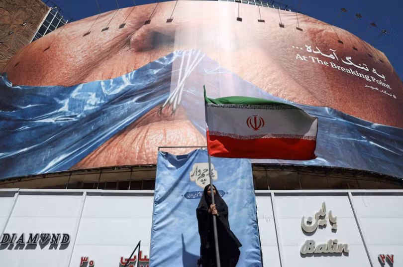 Một người phụ nữ cầm cờ Iran gần một bảng quảng cáo chống Mỹ có hình Tổng thống Mỹ Donald Trump và eo biển Hormuz, tại Tehran, Iran, ngày 25/5. Ảnh: Reuters.
