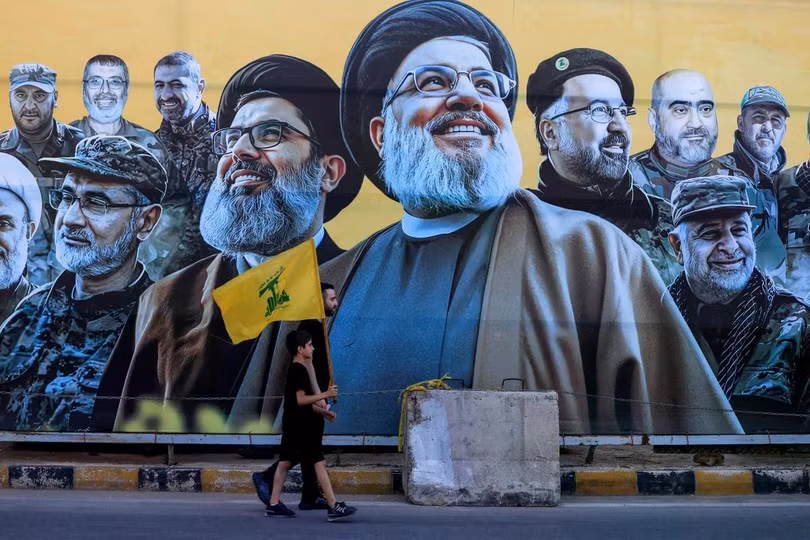 Một người đàn ông dắt một cậu bé cầm cờ Hezbollah đi ngang qua một bức tranh tường bên ngoài Beirut, Lebanon, hôm 17/6. Thỏa thuận giữa Mỹ và Iran bao gồm các điều khoản về lệnh ngừng bắn ngay lập tức và vĩnh viễn trên tất cả các mặt trận, bao gồm cả Lebanon. Ảnh: Reuters.