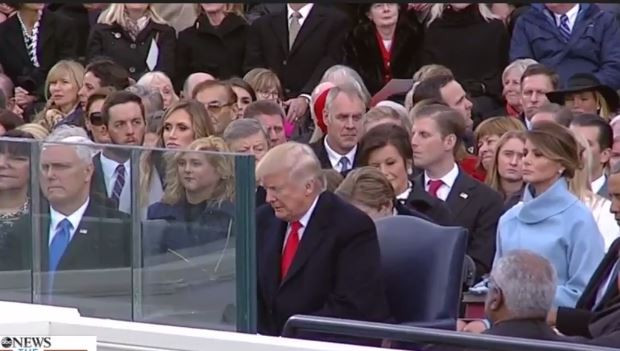 Ông Trump ngồi ở hàng ghế đầu trên lễ đài, phía trước có tấm kính chắn bảo vệ.