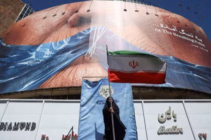 Một người phụ nữ cầm cờ Iran gần một bảng quảng cáo chống Mỹ có hình Tổng thống Mỹ Donald Trump và eo biển Hormuz, tại Tehran, Iran, ngày 25/5. Ảnh: Reuters.